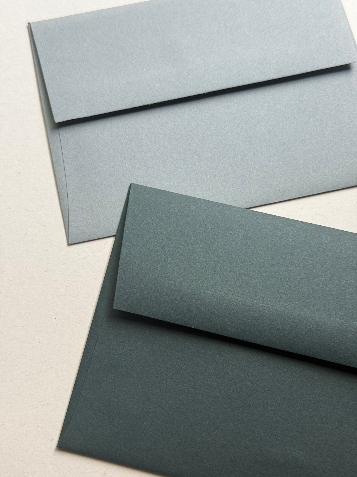 Two textured paper envelopes in different muted blue-gray shades, one light blue-gray and one dark blue-gray, arranged diagonally on a light beige background.
