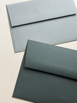 Two textured paper envelopes in different muted blue-gray shades, one light blue-gray and one dark blue-gray, arranged diagonally on a light beige background.