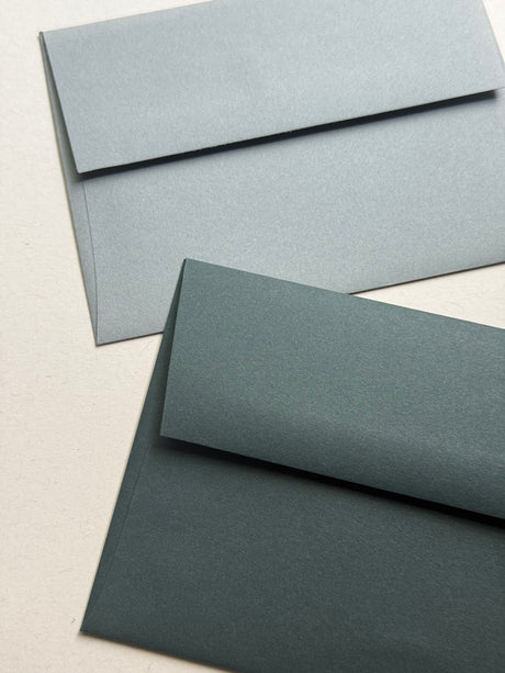 Two textured paper envelopes in different muted blue-gray shades, one light blue-gray and one dark blue-gray, arranged diagonally on a light beige background.